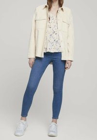 Leichte cremefarbene Knopfjacket über einem blumengemusterten Oberteil, kombiniert mit blauen Skinny-Jeans und weißen Sneakers. Schlichtes Design, lässige Passform.