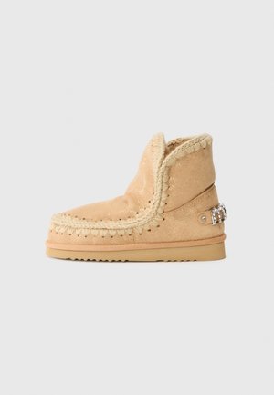 Stivaletti beige alla caviglia con una superficie morbida e luccicante, dettagli intrecciati lungo il bordo e accessori decorativi sul retro. Suola piatta.