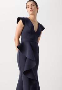 Robe bleu marine avec un décolleté en V profond et des manches courtes. Présente un buste cache-cœur et un accent de volants en cascade à partir de la taille. Texture lisse.
