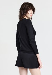 Giovane donna con capelli scuri e mossi che indossa un top nero a maniche lunghe e pantaloni corti neri, mentre guarda oltre la spalla su uno sfondo bianco.
