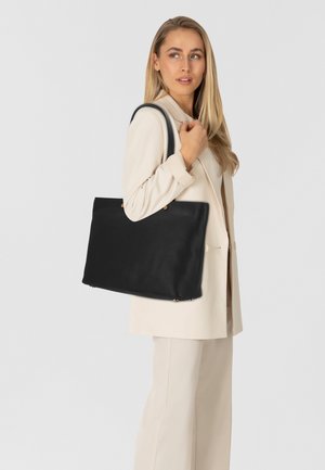 Schwarze Leder-Shopper-Tasche mit einer genarbten Textur, ausgestattet mit zwei Tragegriffen und Metallelementen. Neben einer Person in heller Kleidung dargestellt.
