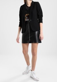 Femme portant un blazer noir, un t-shirt graphique, une mini-jupe en cuir noir avec zip avant, et des baskets montantes blanches sur fond uni.