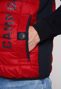 Giacca a vento rossa con dettagli blu navy, dotata di una tasca laterale e un patch con logo. Il materiale sembra imbottito e ha una texture liscia.