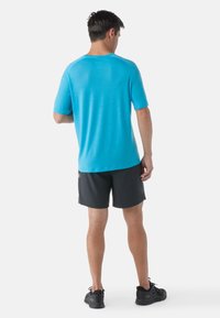 T-shirt de sport turquoise à manches courtes, col rond et texture lisse, associé à un short noir et des chaussures de course.