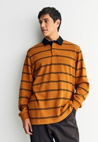 Senf žuta polo majica s dugim rukavima s crnim horizontalnim prugama, sa crnim ovratnikom i trostrukim gumbima. Nošena s tamnim hlačama.