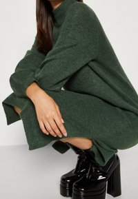 Donna accovacciata che indossa un abito lavorato a maglia verde scuro e stivaletti neri lucidi con tacco a blocco su uno sfondo bianco.