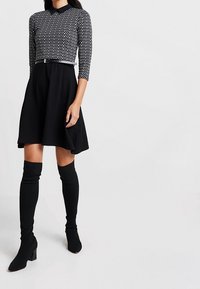 Robe noire avec un corsage ajusté à pois, des manches trois-quarts, une jupe évasée et une fine ceinture à la taille. Bottes noires montant jusqu'aux genoux.