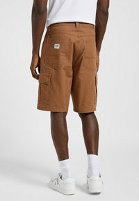 Pantalones cortos de carga marrones confeccionados en algodón, con múltiples bolsillos, costuras reforzadas y un corte recto. Usados con zapatillas deportivas blancas y calcetines.