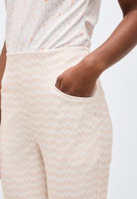 Άτομο που φοράει ανοιχτό μπεζ παντελόνι με σχέδιο και το χέρι του στην μπροστινή τσέπη, συνδυασμένο με λευκό πουκάμισο με μικρές πορτοκαλί λεπτομέρειες.