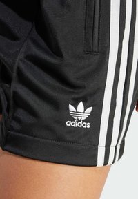 Černé atletické šortky z hladké látky s bílým logem Adidas a třemi svislými bílými pruhy na bocích.