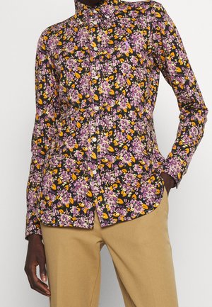 Chemise boutonnée à manches longues avec imprimé floral violet et jaune portée avec un pantalon beige, main du modèle reposant dans la poche du pantalon.