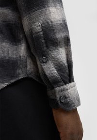 Schwarz-weißer Karomuster-Hemd, strukturiertes Material, Knopfmanschetten, kontrastierende Farben und sichtbare Nähdetails am Saum. Schwarze Unterhose.