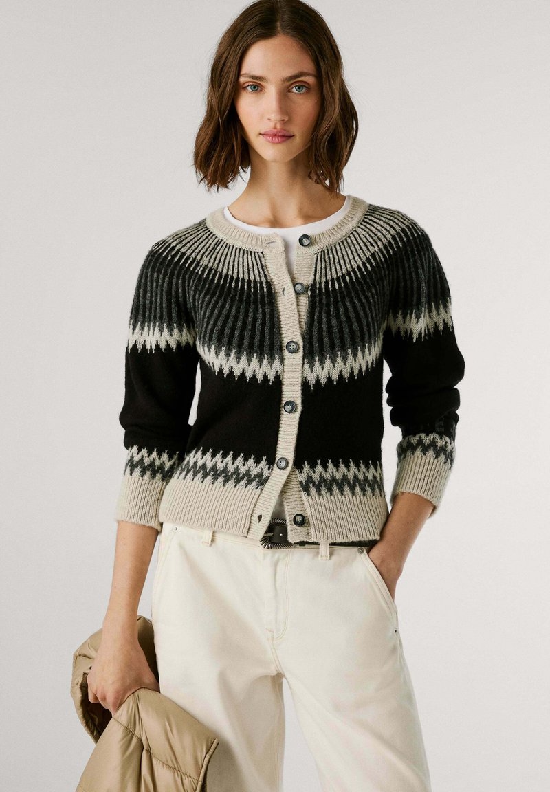 Cárdigan de punto negro y crema con un canesú texturizado, patrón de zigzag en contraste y cierre de botones. Lleva encima una blusa clara con pantalones beige.