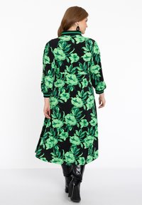Blumenkleid mit grünen Blumen und Blättern auf schwarzem Hintergrund, mit 3/4 Ärmeln und einem plissierten Saum, kombiniert mit schwarzen knielangen Stiefeln.
