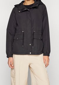 Giacca nera con cappuccio, polsini elastici e orlo regolabile con coulisse, dotata di due tasche frontali e bottoni a pressione. Tessuto liscio con finitura opaca.