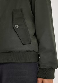 Veste bomber vert olive avec un ourlet et des poignets côtelés, dotée d'une grande poche à rabat avec un bouton en métal. Texture de tissu lisse et léger.