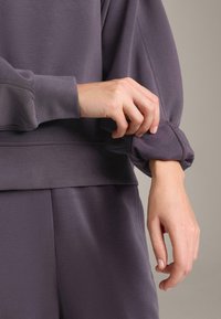 Maglietta a maniche lunghe viola con dettaglio a coste ai polsini e tessuto liscio. La manica è parzialmente arrotolata, rivelando una silhouette aderente.