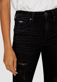 Vaqueros de mezclilla negros de cintura alta con un diseño desgastado, presentando roturas en las rodillas y un suave brillo. Acompañados de una blusa ajustada de manga corta blanca.