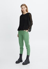Zwart geplisseerde top met lange mouwen, gecombineerd met groene op maat gemaakte broek en zwarte veterschoenen. De outfit heeft een gestructureerde stof en een losse pasvorm.