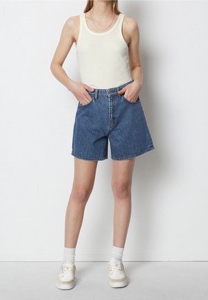 Femme portant un haut blanc sans manches, un short en denim bleu, des chaussettes blanches et des baskets blanches, debout les mains dans les poches contre un mur blanc.