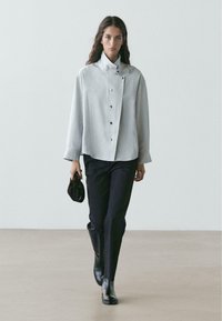 Giacca grigia chiaro con bottoni e collo alto, abbinata a pantaloni neri e stivaletti neri eleganti. La modella porta una piccola borsa scura.