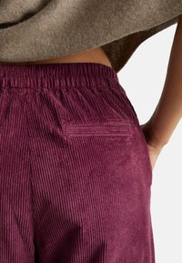 Close-up of a person wearing plum-colored corduroy pants with an elastic waistband and a back welt pocket, paired with a beige sweater.