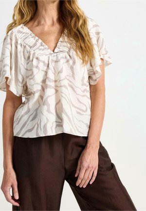 Kobieta ubrana w białą, luźną bluzkę z krótkim rękawem, ozdobioną delikatnym wzorem zebry, oraz ciemnobrązowe szerokie spodnie na tle jednolitego tła.