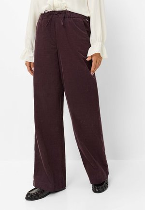 Vrouw draagt losse donkerpaarse ribfluwelen broek met koord in de taille, gecombineerd met een witte blouse met lange mouwen en zwarte platte schoenen.