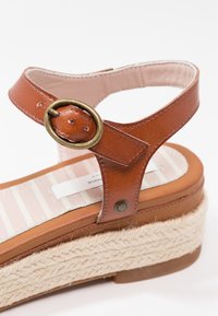 Sandalo in pelle marrone con una fibbia metallica circolare, piattaforma in juta intrecciata e soletta a righe. Presenta un tacco piatto e design con cinturino alla caviglia.
