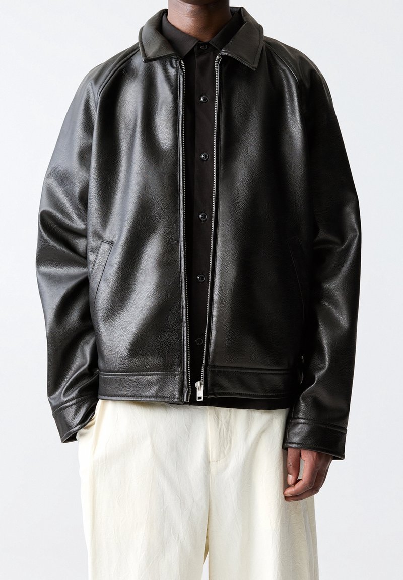 Homme portant une veste zippée en cuir noir sur une chemise noire boutonnée et un pantalon blanc ample sur un fond uni.