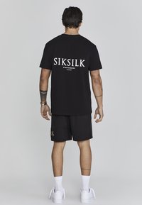 Black short-sleeve t-shirt with white "SIKSILK BY INVITATION ONLY" text on the back. Paired with black shorts featuring gold "SIKSILK" text.