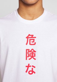 Osoba v bílém tričku s vertikálními červenými japonskými znaky uprostřed, které říkají "nebezpečí" nebo "hazard."