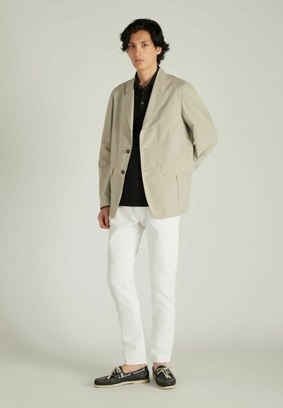 Chaqueta beige con un diseño entallado, cierre de botones y dos bolsillos frontales, combinada con un polo negro, pantalones blancos y zapatos negros.