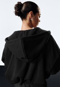 Crna oversized majica s kapuljačom s mekanim, teksturiranim unutarnjim slojem i dubokom kapuljačom, s opuštenim krojem i suptilnim detaljima šavova.