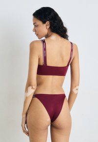 Burgunder bikini-set som har en tettsittende topp med justerbare stropper og matchende lavt belte truser, begge med logo-detaljer på stroppene.