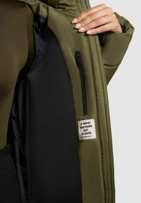 Groene geïsoleerde jas met een ritszak, zwarte voering en een stoffen label met tekst. De buitenkant heeft een gladde, matte textuur.