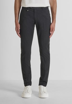 Hombre con pantalones negros ajustados y zapatillas blancas, de pie frente a un fondo claro y liso.