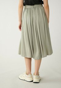 Jupe vert olive clair, longueur genou, avec des plis et une taille élastique, assortie à des baskets blanches et beiges. Texture lisse.
