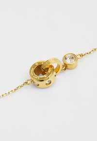 Pulseira de ouro com um pendente circular com texto gravado e cristais transparentes embutidos. Design de corrente com um pequeno acento circular.