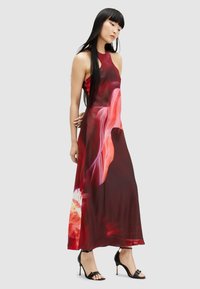 Maxi dress featuring a halter neck, red and pink abstract patterns, made from smooth fabric. Paired with black high-heeled sandals.
