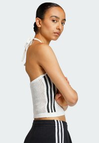 Žena s vlasy staženými dozadu, oblečená v bílém pleteném halterneku s černými pruhy a černých kalhotách s bílými pruhy, stojící se založenýma rukama.