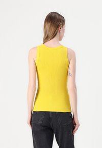Gul ribbet ermeless topp med rund halslinning, tettsittende design og myk tekstur, parret med mørk denim bukser.
