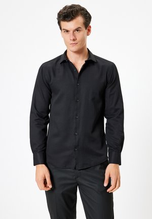 Chemise noire à manches longues avec un col boutonné, présentant une texture lisse et un motif ribbé subtil, assortie à un pantalon sombre.