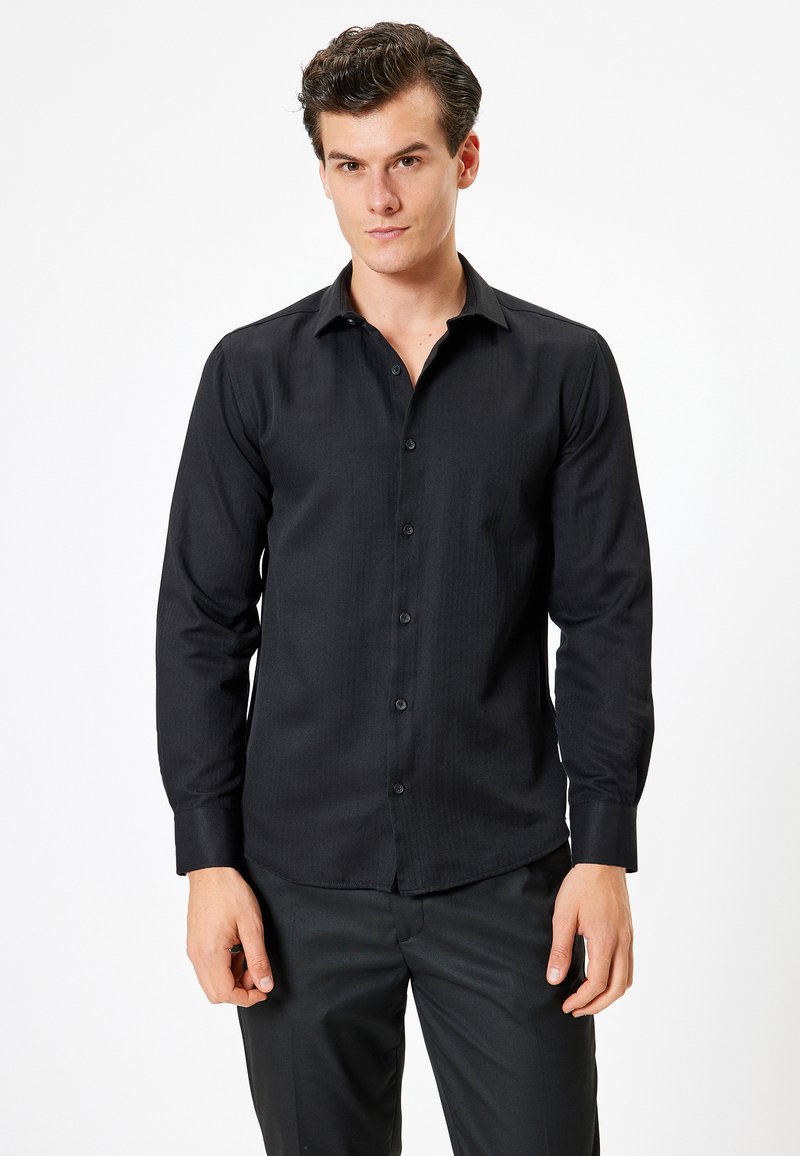 Camicia nera a maniche lunghe con colletto a bottoni, caratterizzata da una texture liscia e un sottile motivo a coste, abbinata a pantaloni scuri.