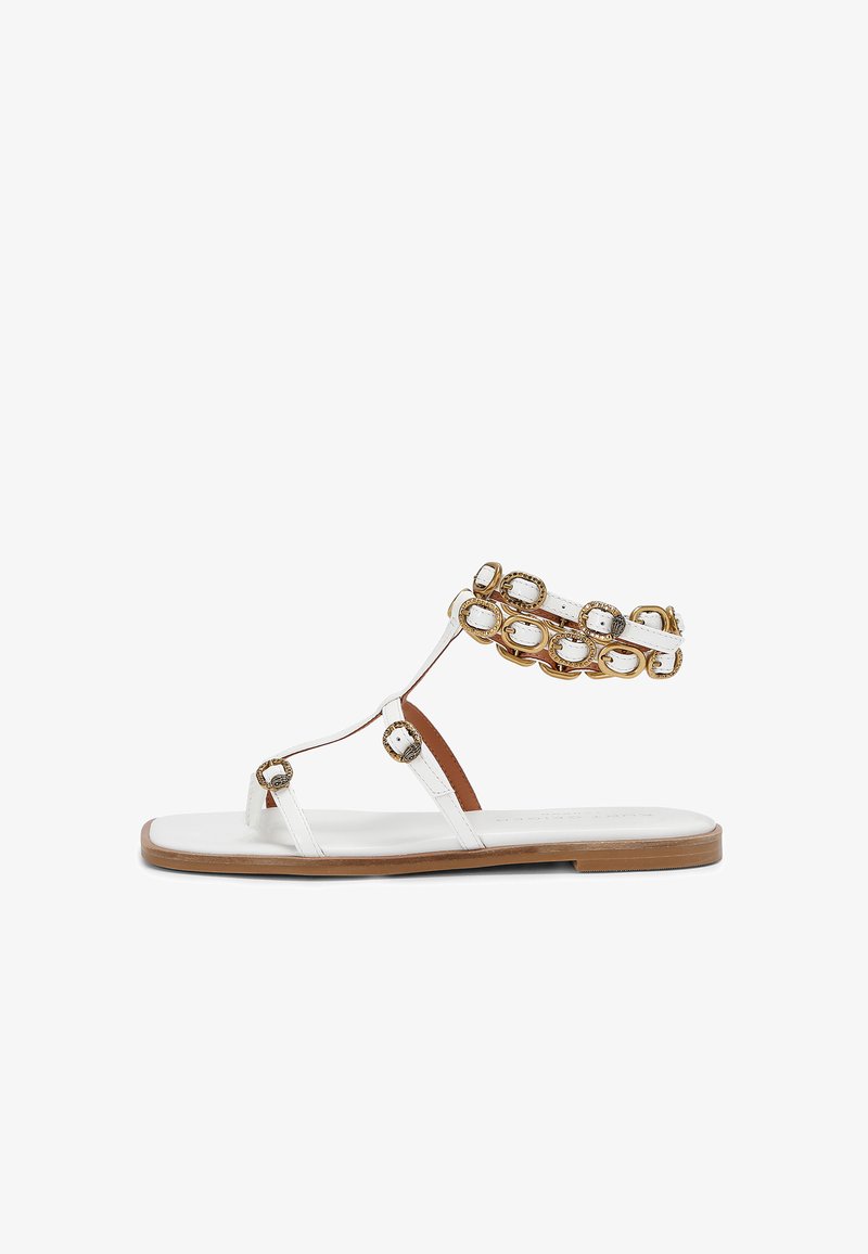 Sandalo piatto bianco con una accentuazione marrone, caratterizzato da una catena decorativa dorata sulla cinghia alla caviglia e da cinghie minimali che collegano alla suola.