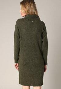Femme aux cheveux blonds portant une longue robe pull en maille vert olive, de dos face à un fond uni.