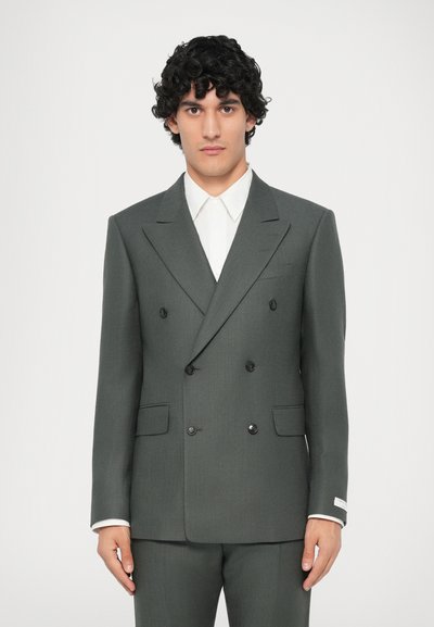 Blazer grigio doppiopetto con tessuto intrecciato, dotato di bottoni neri, revers a lancia e camicia bianca sotto. Vestibilità sartoriale.