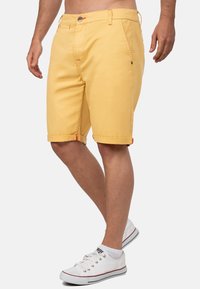 Gele katoenen shorts met opgerolde zoom en rode accenten. Beschikt over zakken en een knoopsluiting. Gekozen in combinatie met witte sneakers.