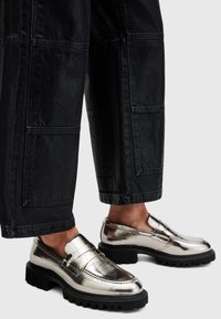 Loaferi argintii metalici cu o talpă groasă neagră, având o textură netedă și un design minimalist. Purtați cu pantaloni negri, largi.