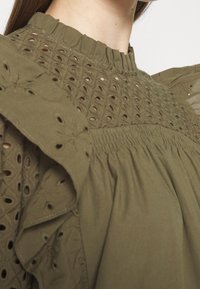 Haut vert olive avec des motifs découpés complexes, bords volantés et texture douce. L'encolure présente un détail froncé.